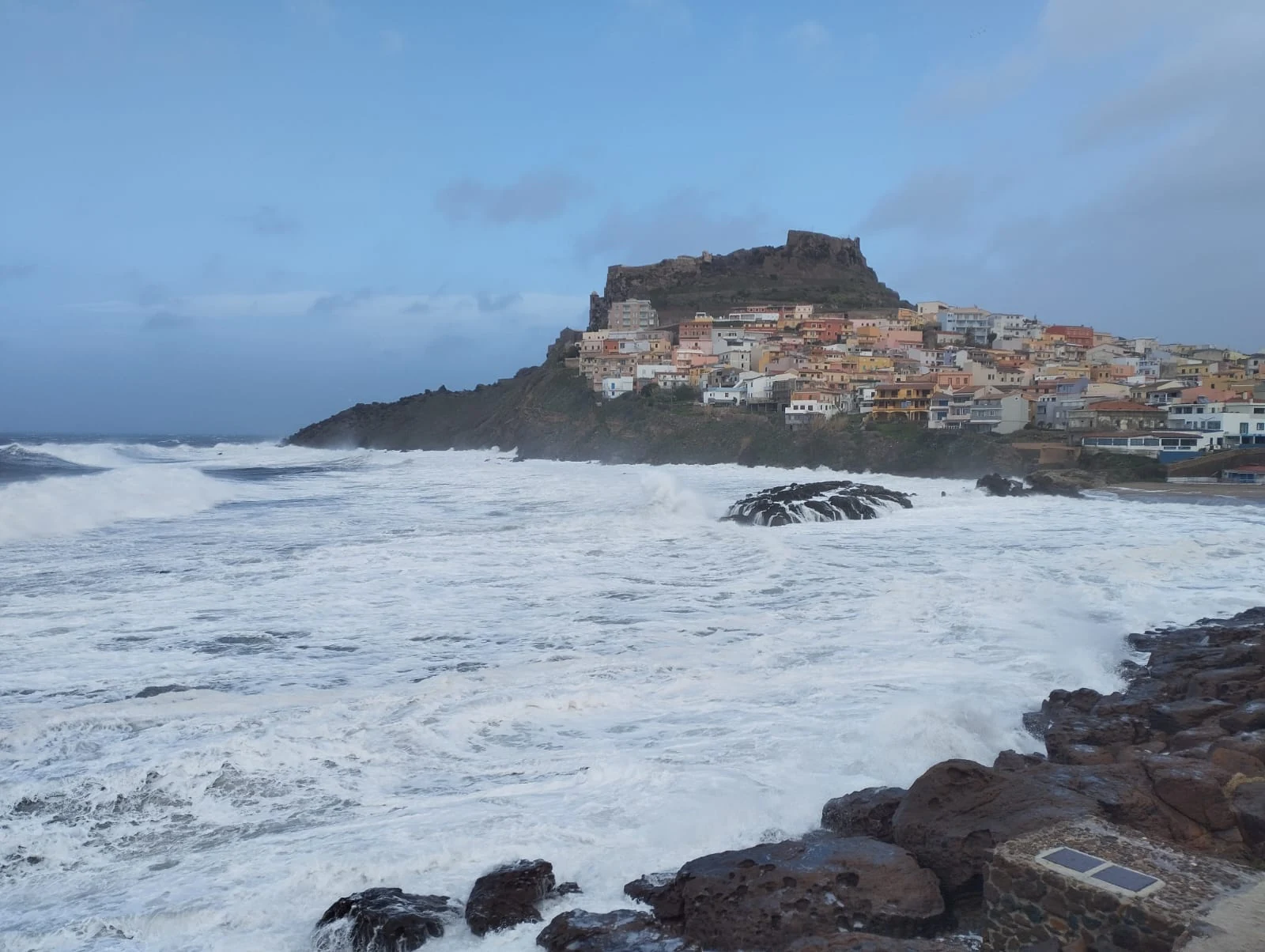 Le onde di una mareggiata sugli scogli, con il borgo di Castelsardo sullo sfondo